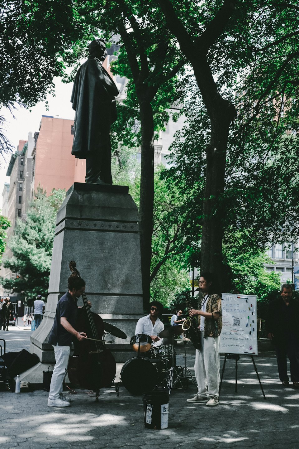 Des musiciens jouent du jazz dans un parc avec une statue et un tableau blanc.